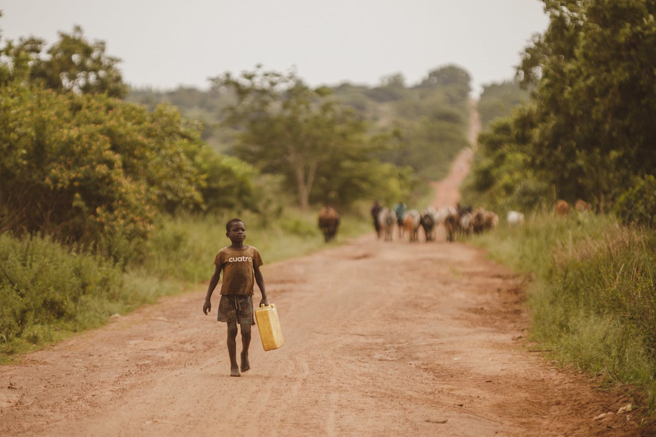 A young boy carrying a yellow container walks along a dirt road in rural Uganda, surrounded by lush greenery.